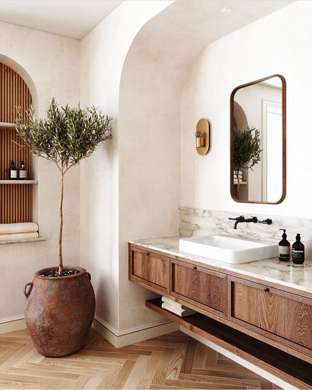 Warm oak bathroom vanity with marble top and brass mirror in an arched alcove by Buxleys